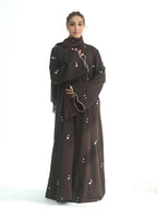 A woman modeling a dark brown abaya with matching hijab and wide sleeves, decorated with small white floral embroidery, set against a plain white background.