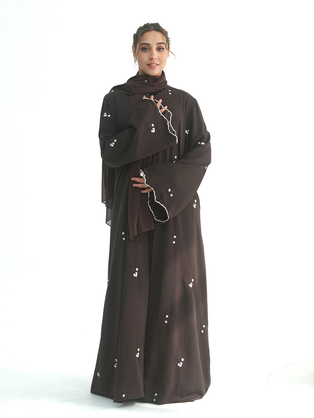 A woman modeling a dark brown abaya with matching hijab and wide sleeves, decorated with small white floral embroidery, set against a plain white background.