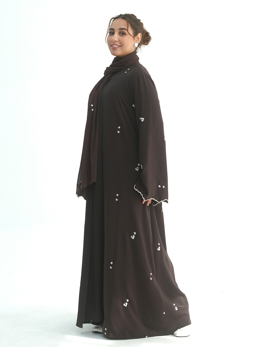 A woman modeling a dark brown abaya with small silver floral embroidery, shown from a side profile against a solid white studio background.