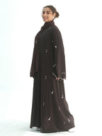 A woman modeling a dark brown abaya with small silver floral embroidery, shown from a side profile against a solid white studio background.
