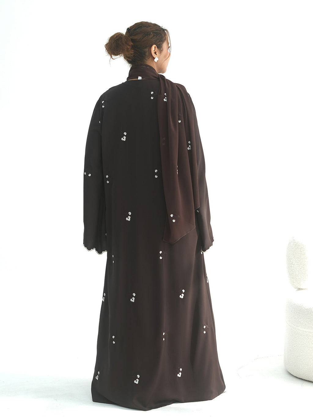 A woman viewed from behind wearing a long dark brown abaya with small white embroidered details and a matching draped scarf, set against a plain white studio background.