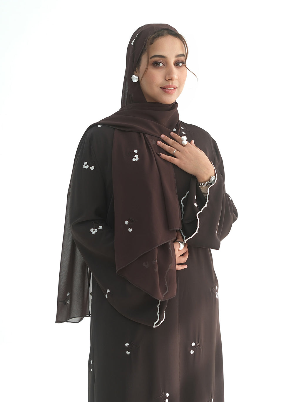 A woman wearing a dark brown abaya with small white embroidered details, a matching brown hijab, and pearl earrings, posed against a solid white background.