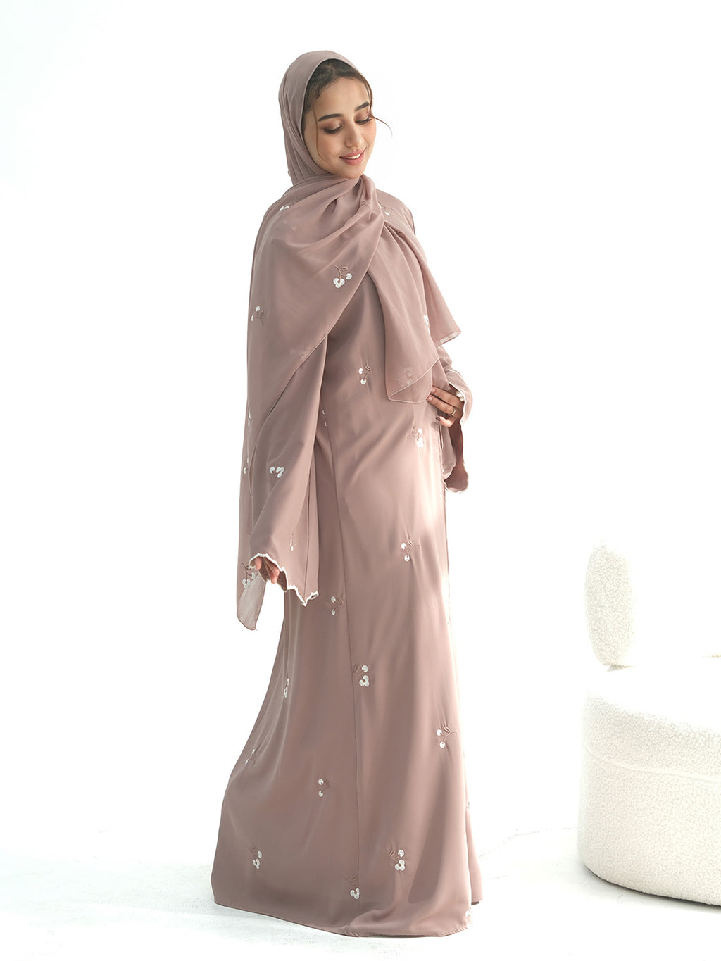 A woman wearing a modest dusty rose abaya and matching hijab, standing against a bright white studio background.