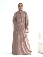 A woman wearing a long-sleeved, floor-length mauve-brown abaya with small white embroidered details, a matching hijab, and white slip-on shoes, standing against a plain white studio background.