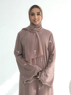 A woman wearing a matching modest outfit in a dusty mauve color, including a long-sleeved abaya with white floral embroidery and scalloped edges, a coordinating hijab, and a long scarf, set against a plain white studio background.