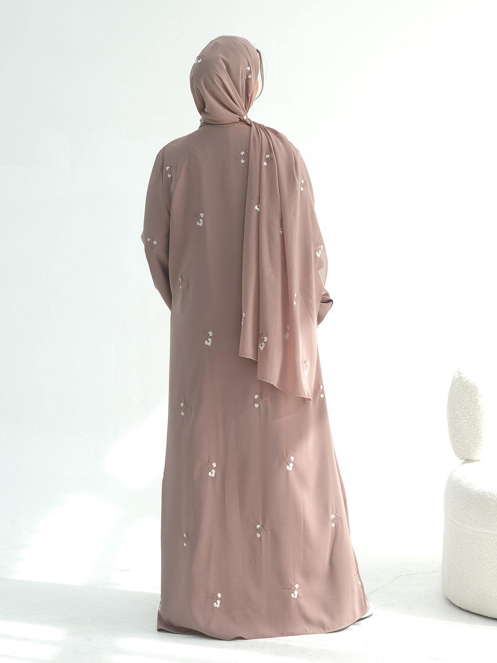 A woman wearing a dusty rose abaya and matching hijab, shown from the back. The abaya features small white embroidered details scattered across the fabric.