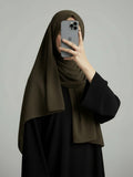 A woman wearing a dark olive green hijab and a black abaya takes a mirror selfie with her phone against a plain gray background.