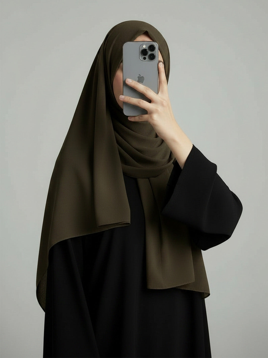 A woman wearing a dark olive green hijab and a black abaya takes a mirror selfie with her phone against a plain gray background.