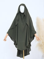 An olive green modest drape or chador with a hood and wide draped sleeves, displayed on a mannequin against a light background with a decorative pampas grass prop.