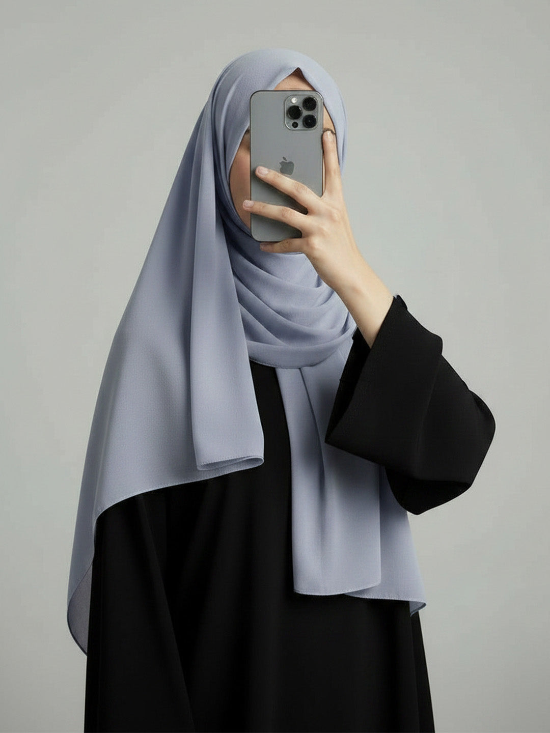 A woman wearing a light gray hijab and a black abaya takes a mirror selfie with her phone against a plain gray background.