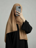 A woman wearing a beige hijab and a black abaya takes a mirror selfie with an iPhone against a plain gray studio background.