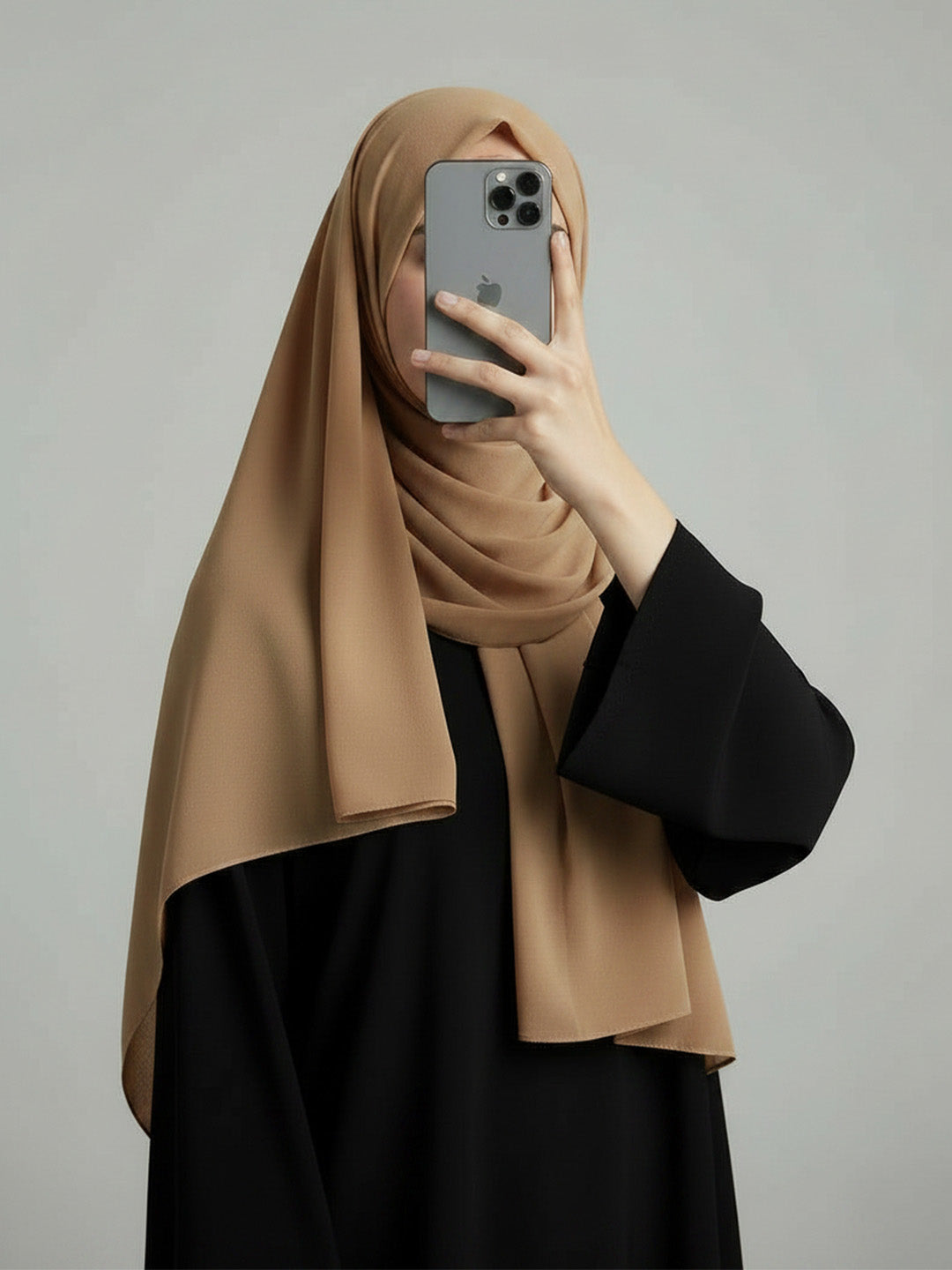 A woman wearing a beige hijab and a black abaya takes a mirror selfie with an iPhone against a plain gray studio background.