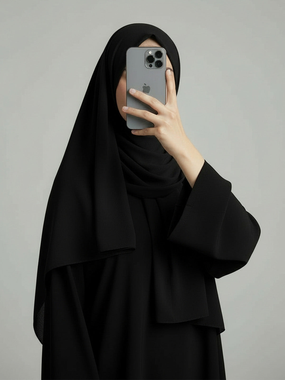 A person wearing a black hijab and a matching black abaya takes a mirror selfie with a gray smartphone against a plain light gray background.