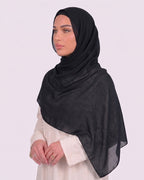 Model wearing a lightweight, black chiffon hijab wrapped elegantly over a beige long-sleeved dress.
