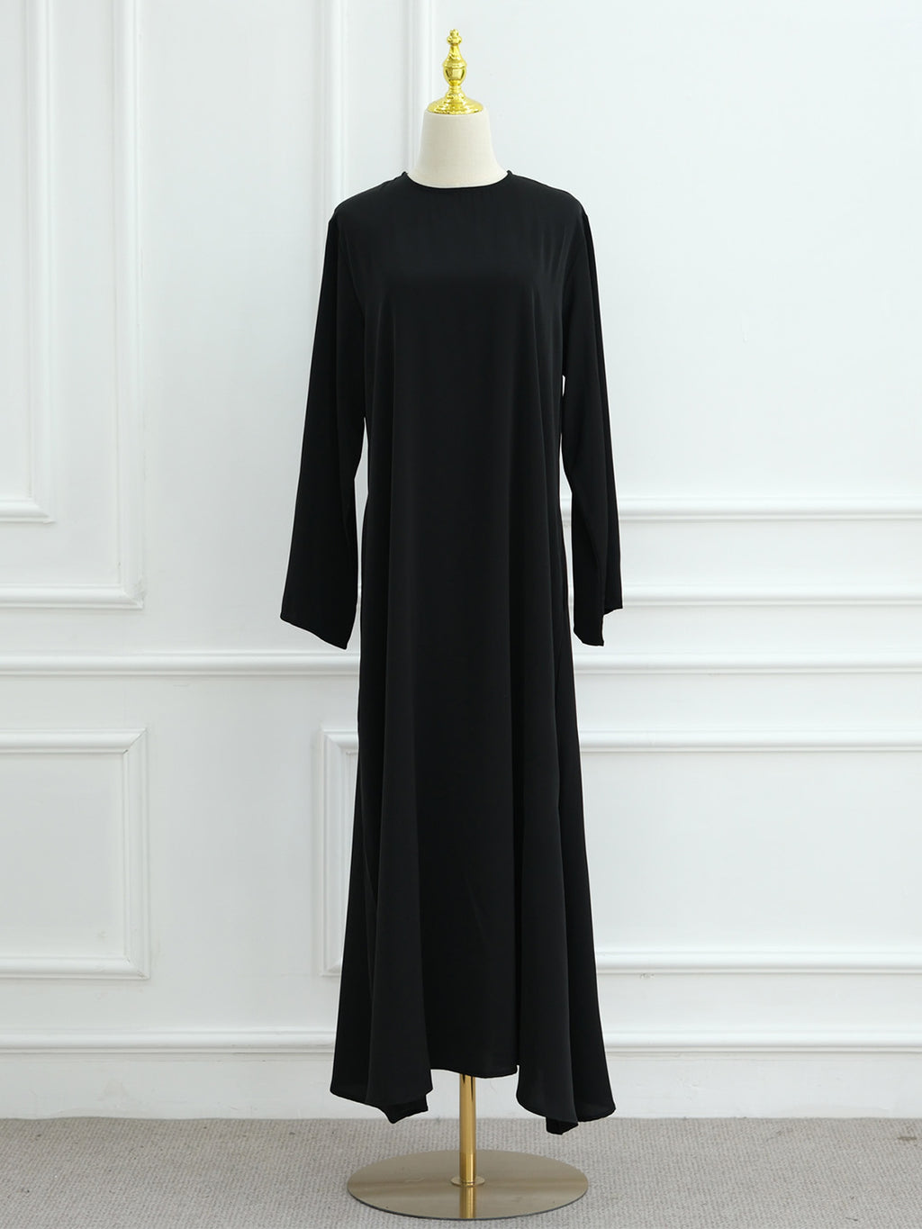 A long-sleeve black maxi dress displayed on a cream dress form with a gold finial and stand, set against a white paneled wall.