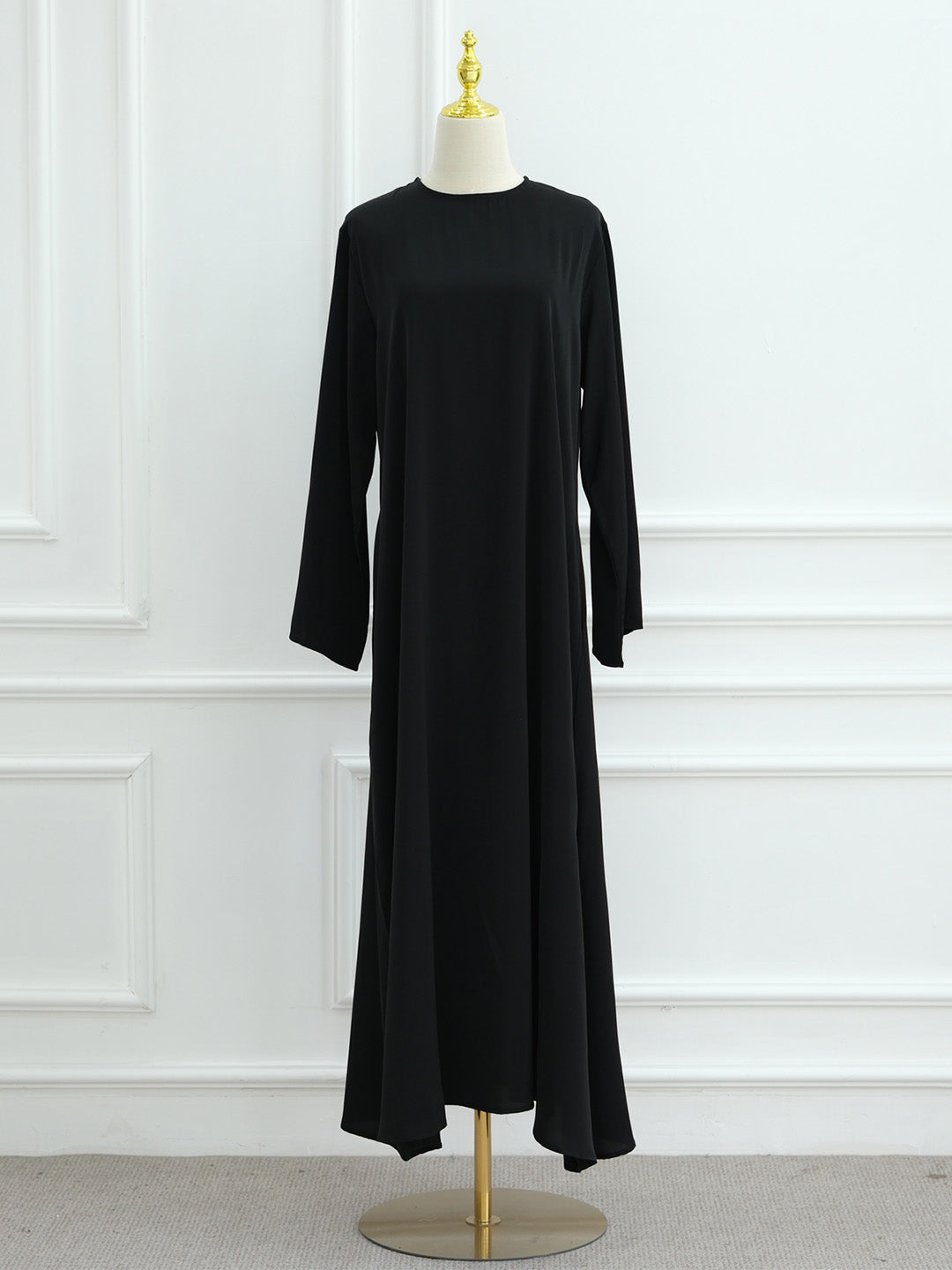 A long-sleeve black maxi dress displayed on a cream dress form with a gold finial and stand, set against a white paneled wall.