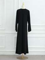 A long black dress displayed on a mannequin in a studio setting with a white paneled wall and light gray carpet. The mannequin has a gold finial on its neck and a gold stand base.