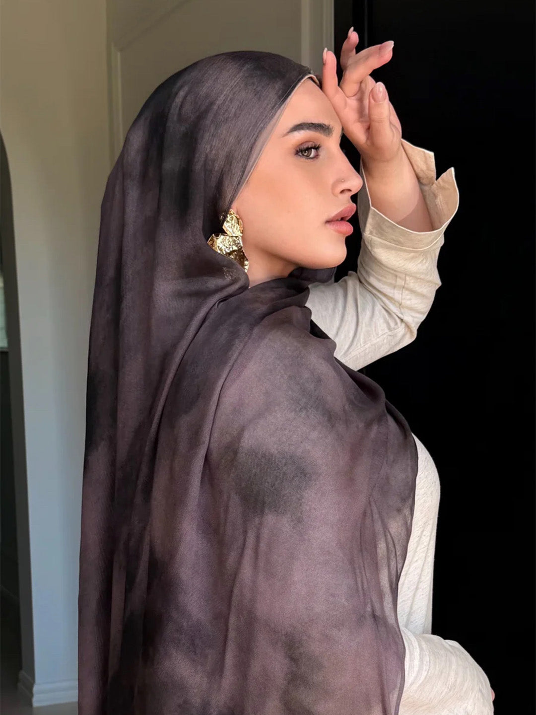 A woman in profile wearing a sheer, dark plum hijab and a light beige long-sleeve top, with a large gold earring visible.