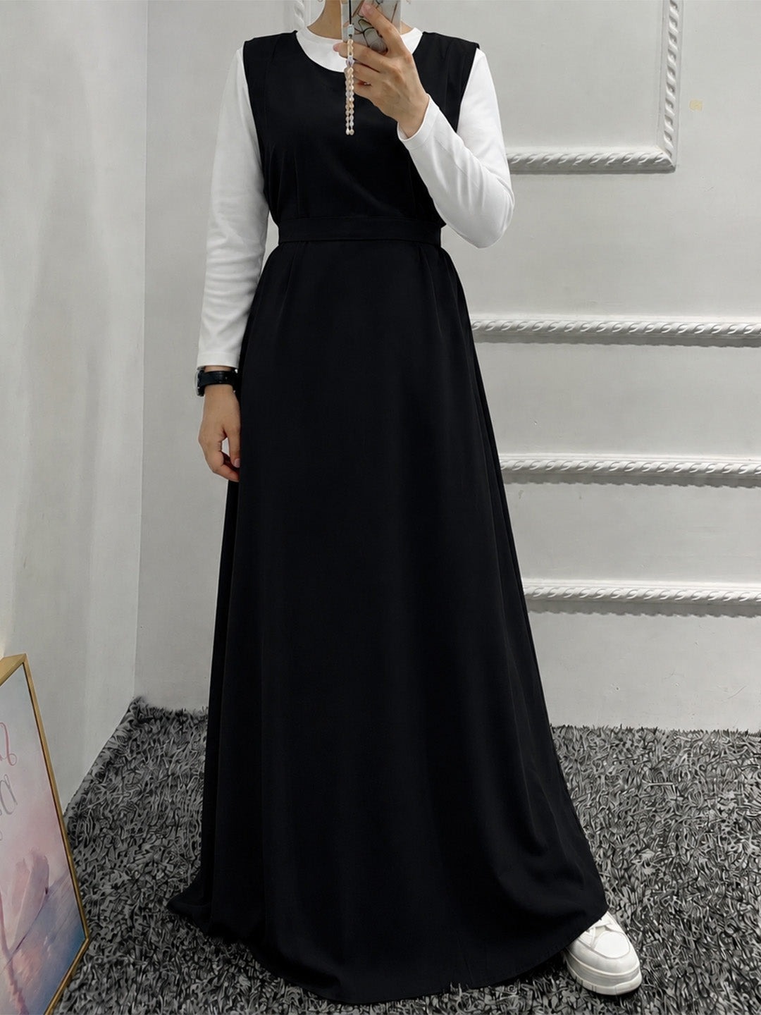 A woman modeling a long black dress with a white long-sleeve collar, paired with white sneakers, standing in an indoor setting with a gray carpet and white paneled walls.