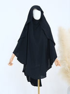 A black modest garment with a draped hood and wide flowing sleeves, displayed on a mannequin against a light background with a decorative pampas grass prop.