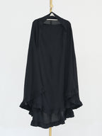 A black long-sleeve draped top with layered ruffle hems, hanging on a black hanger from a gold-colored stand against a plain white background.
