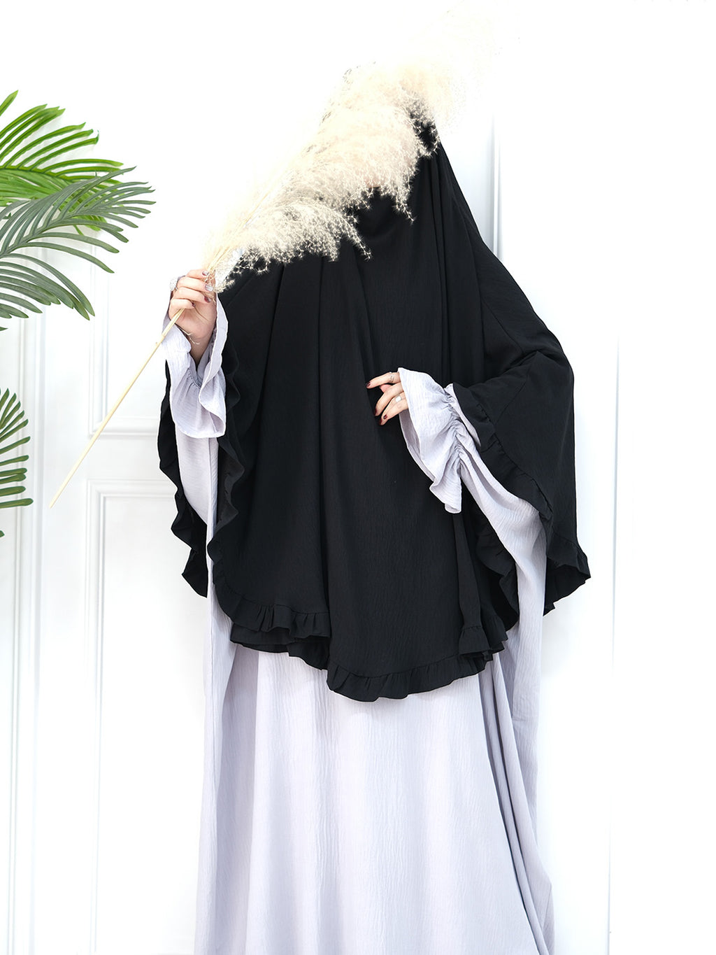 A person wearing a black full-length abaya with wide, layered sleeves and a matching black head covering, styled over a light gray dress. They are holding a decorative pampas grass stem against a white paneled door background with a green plant leaf visible on the left.
