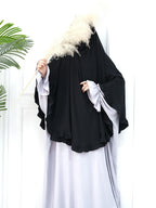 A person wearing a black full-length abaya with wide, layered sleeves and a matching black head covering, styled over a light gray dress. They are holding a decorative pampas grass stem against a white paneled door background with a green plant leaf visible on the left.