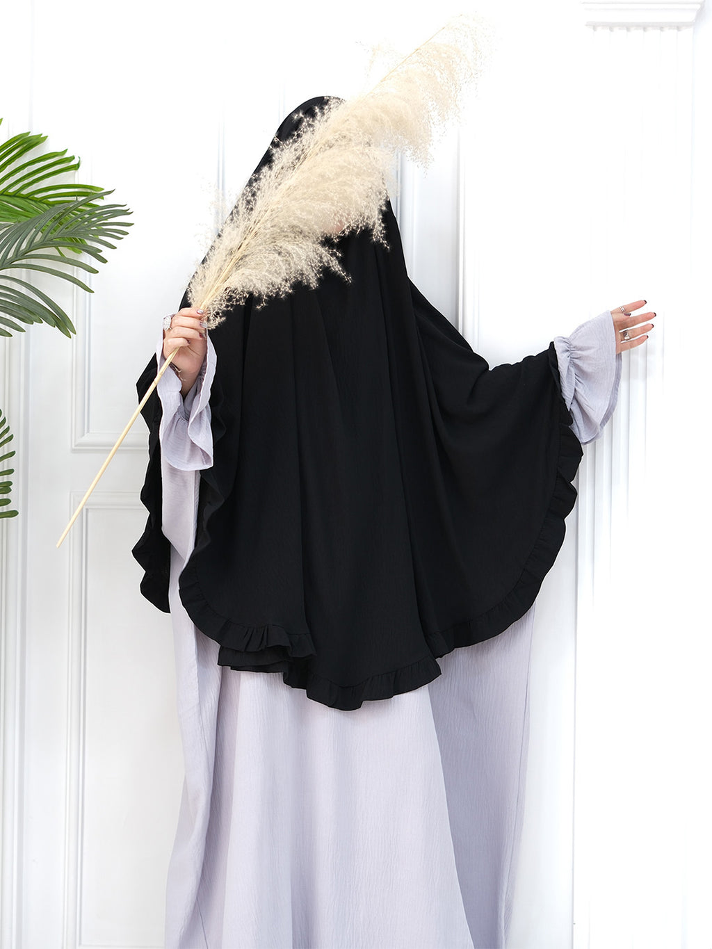 A woman wearing a black full-length modest garment with wide draped sleeves and a light lavender abaya, standing against a white paneled wall and holding a large beige pampas grass stem.