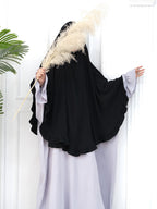A woman wearing a black full-length modest garment with wide draped sleeves and a light lavender abaya, standing against a white paneled wall and holding a large beige pampas grass stem.