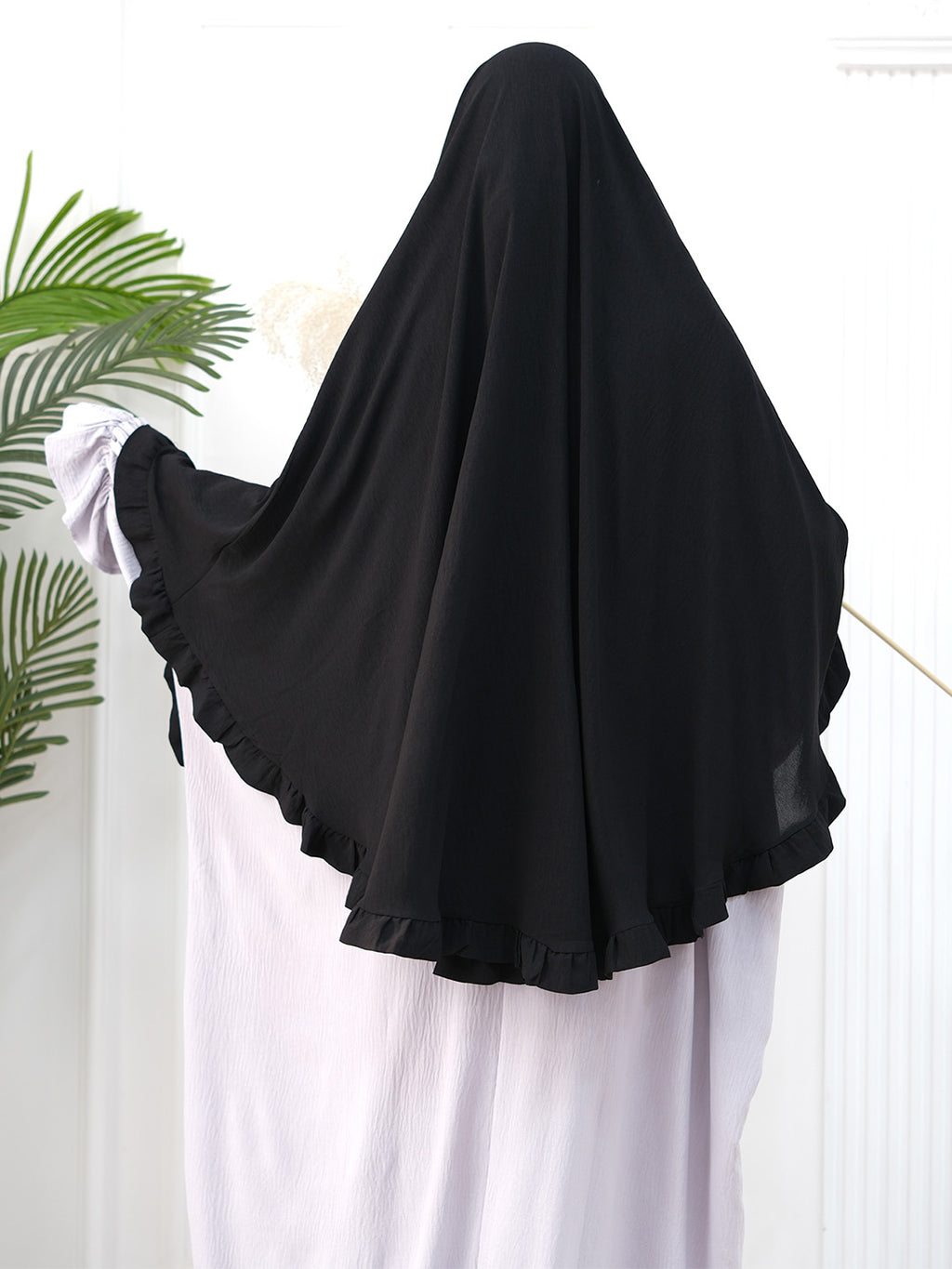 A black full-body modest garment or burkini with wide ruffled sleeves, shown on a mannequin or person from the back against a white indoor background with a plant leaf at the left edge.