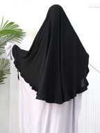 A black full-body modest garment or burkini with wide ruffled sleeves, shown on a mannequin or person from the back against a white indoor background with a plant leaf at the left edge.