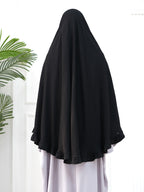 A black modest head covering or burkini draped over a person, shown from the back against a bright white indoor background with a green plant leaf visible on the left.