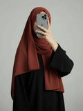 A woman wearing a rust-brown hijab and a black abaya takes a mirror selfie with her phone against a plain gray background.