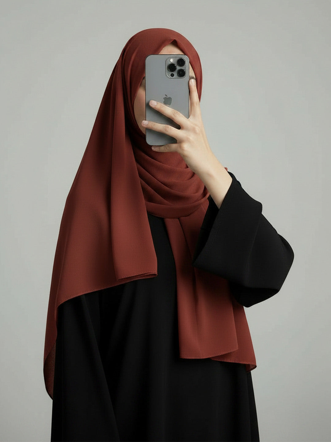 A woman wearing a rust-brown hijab and a black abaya takes a mirror selfie with her phone against a plain gray background.