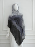 A lightweight hijab scarf with a watercolor-style gradient pattern in shades of gray, charcoal, and black, draped over a white mannequin dress against a white paneled wall.