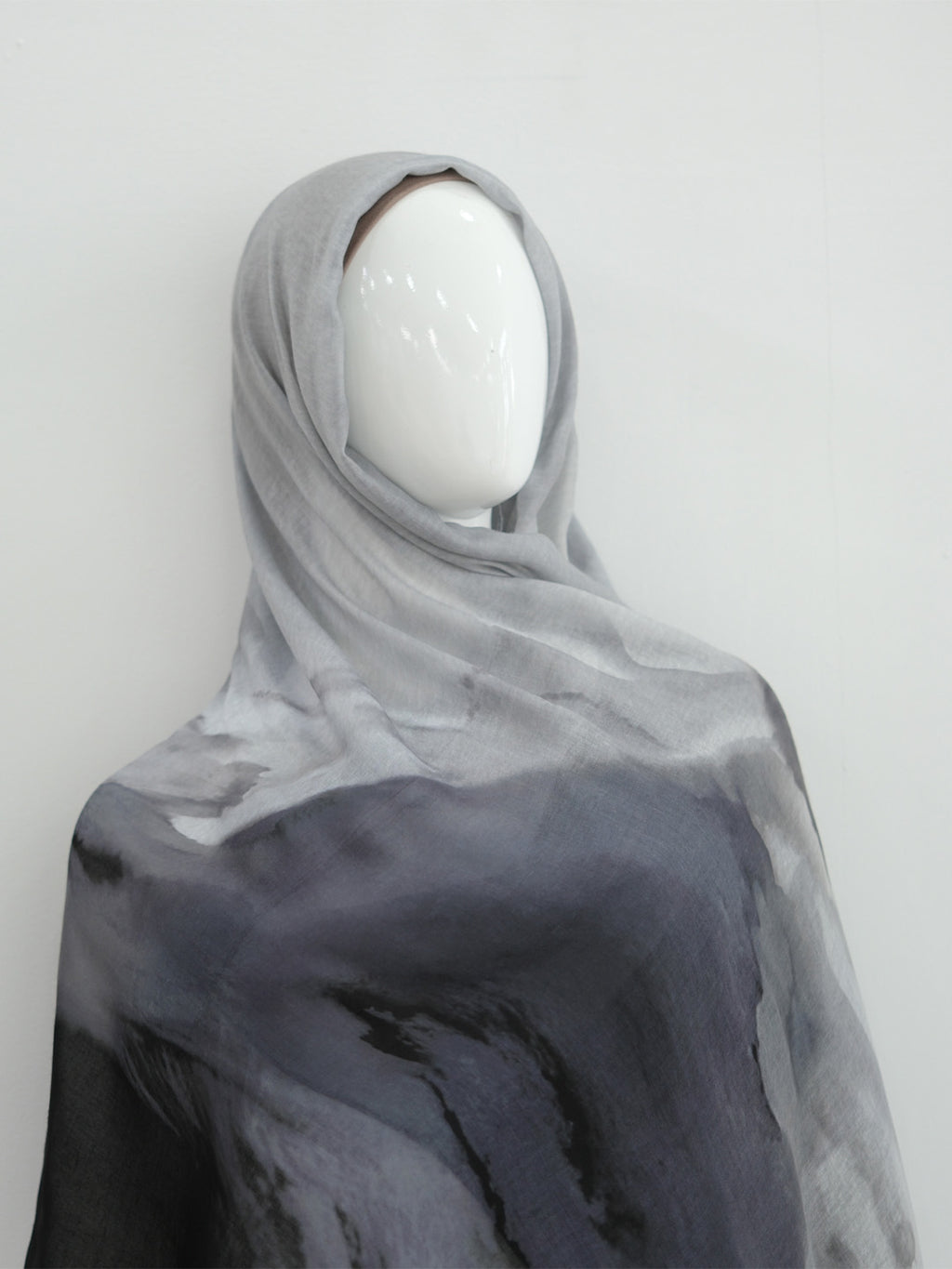A lightweight sheer hijab or shawl draped on a white mannequin head, featuring a watercolor-like gradient from light gray at the top to dark charcoal gray at the bottom.
