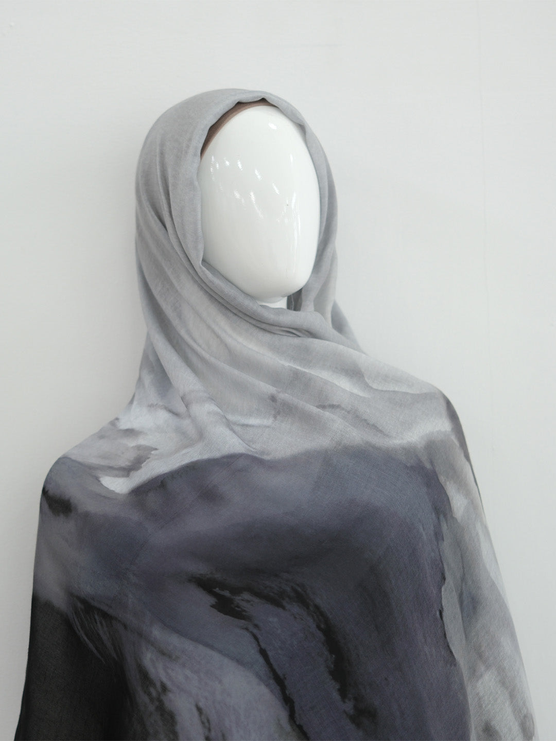 A lightweight sheer hijab or shawl draped on a white mannequin head, featuring a watercolor-like gradient from light gray at the top to dark charcoal gray at the bottom.