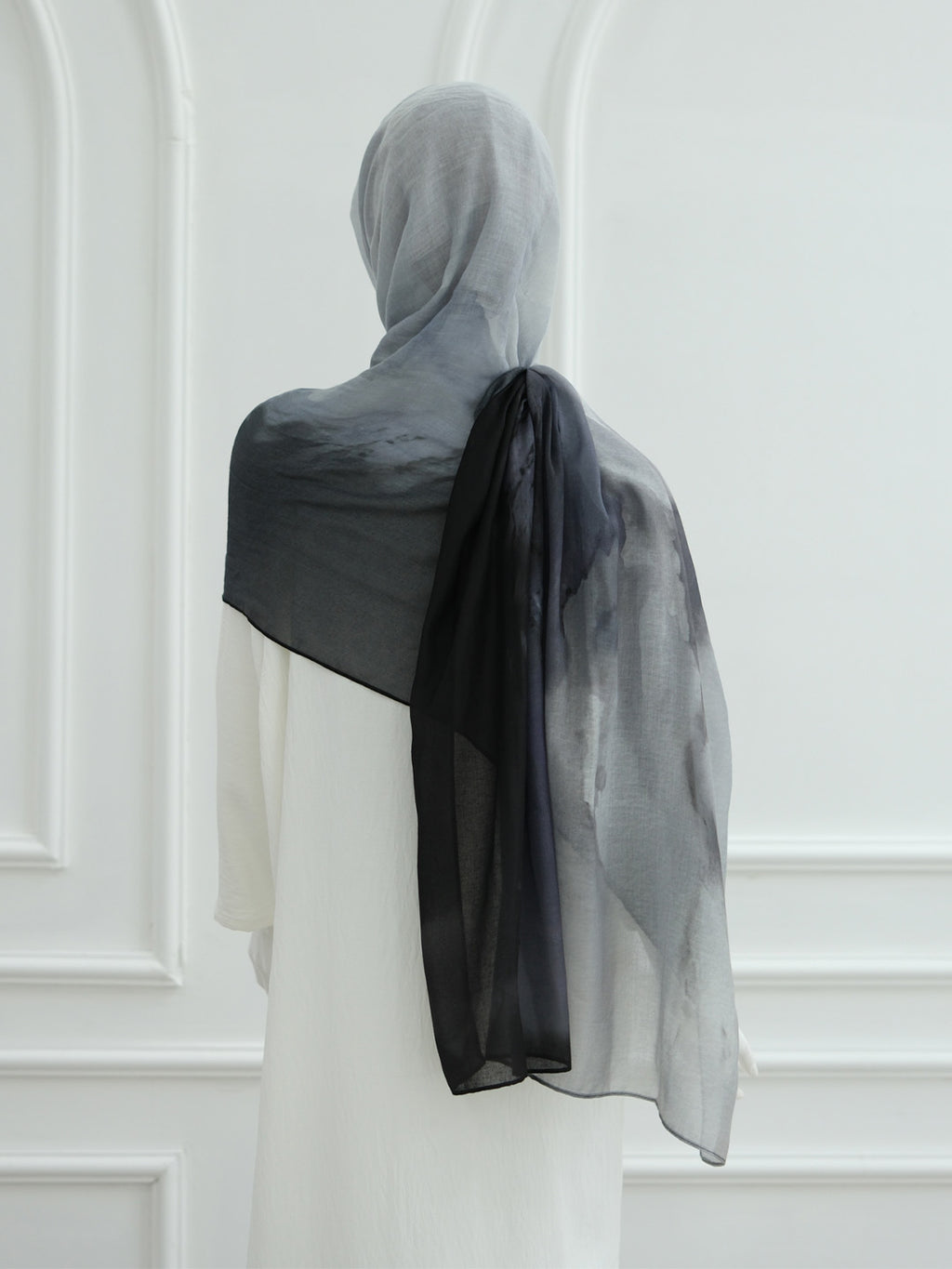 A sheer black and gray ombre hijab draped over the shoulders of a model wearing a white dress, shown from the back against a white paneled wall.