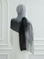 A sheer black and gray ombre hijab draped over the shoulders of a model wearing a white dress, shown from the back against a white paneled wall.