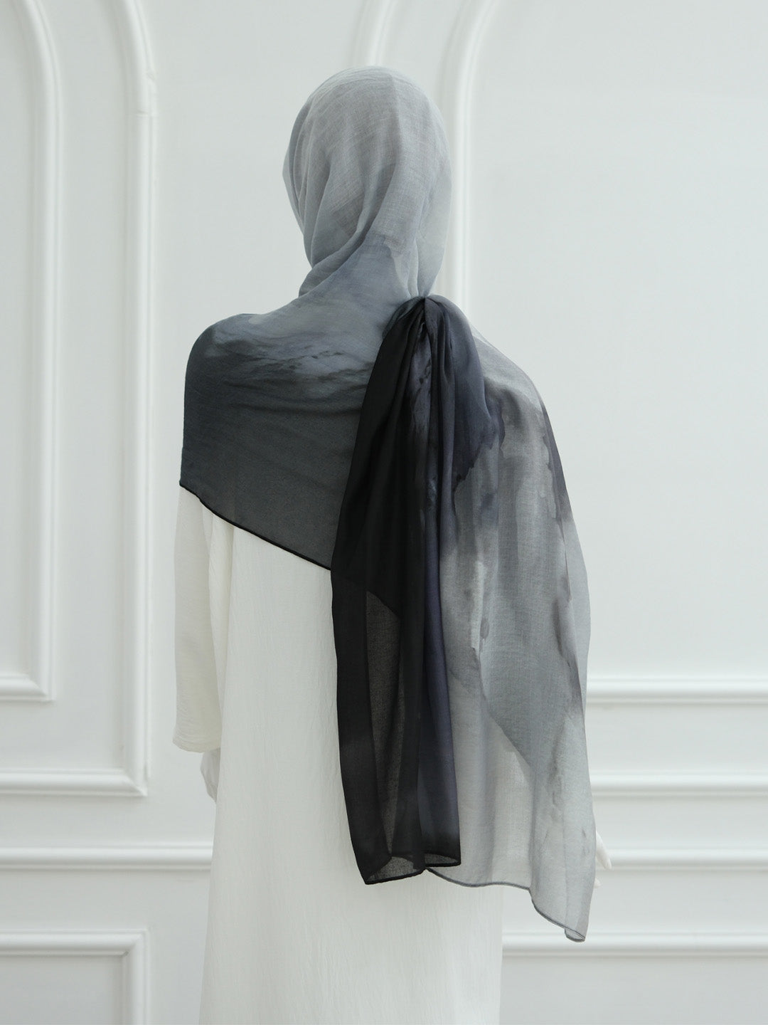 A sheer black and gray ombre hijab draped over the shoulders of a model wearing a white dress, shown from the back against a white paneled wall.