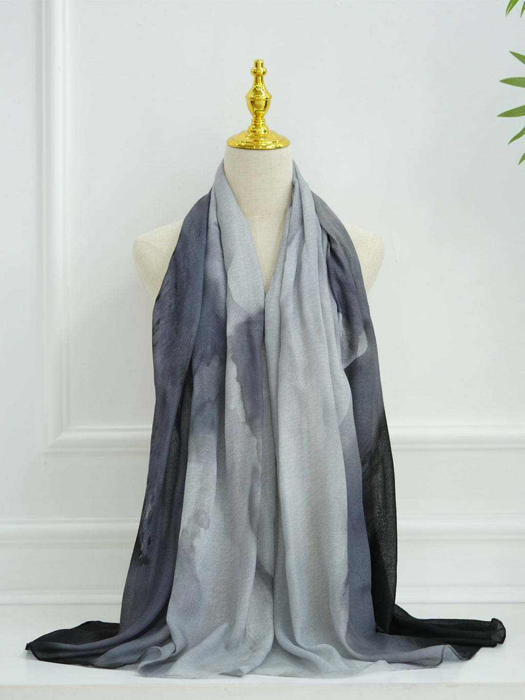 A lightweight gradient scarf draped on a cream-colored mannequin torso with a gold finial, set against a white paneled wall background.