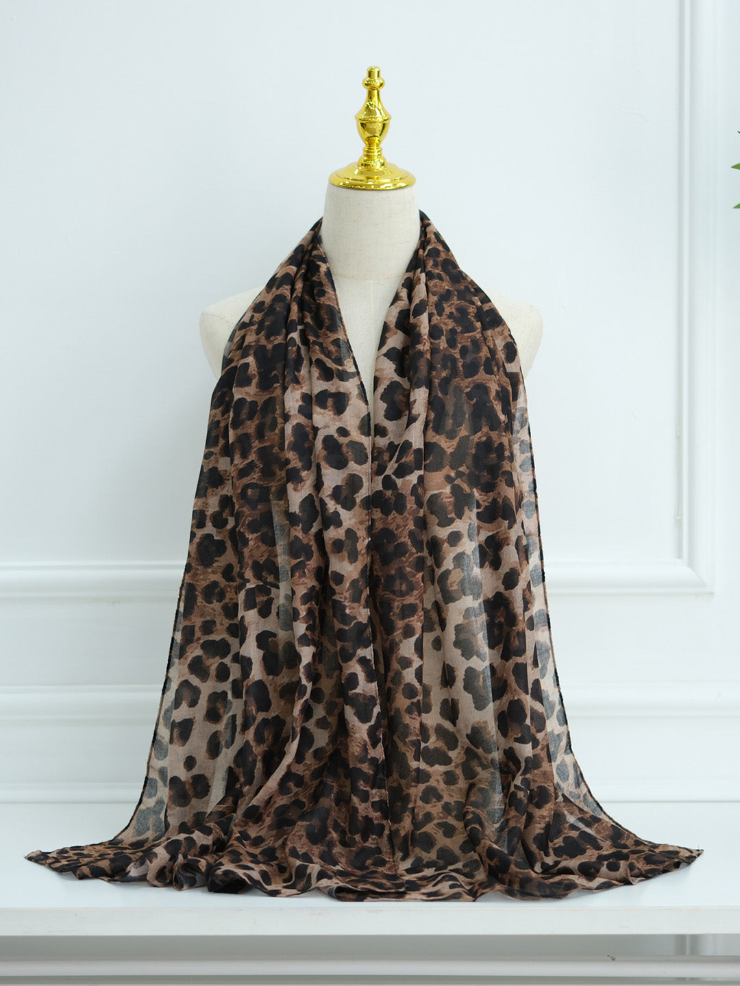 A leopard print scarf draped on a cream mannequin torso with a gold finial, set against a white paneled wall.