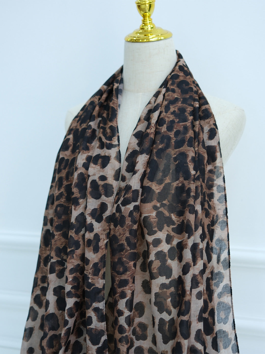 A leopard print scarf draped on a cream-colored mannequin torso with a gold neck cap, set against a light blue wall.