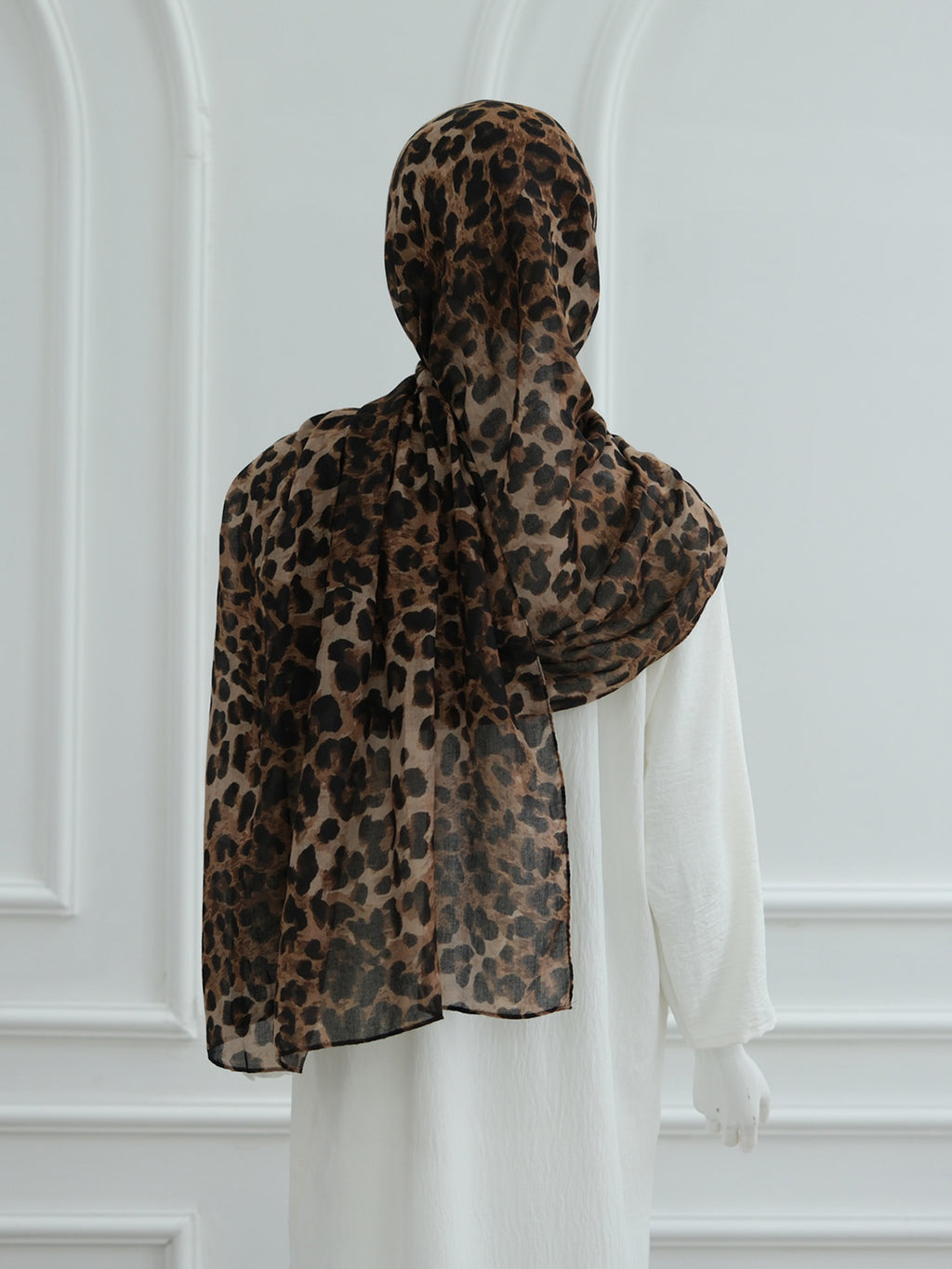 A leopard print hijab draped over a mannequin wearing a white dress, shown from the back against a white paneled wall.
