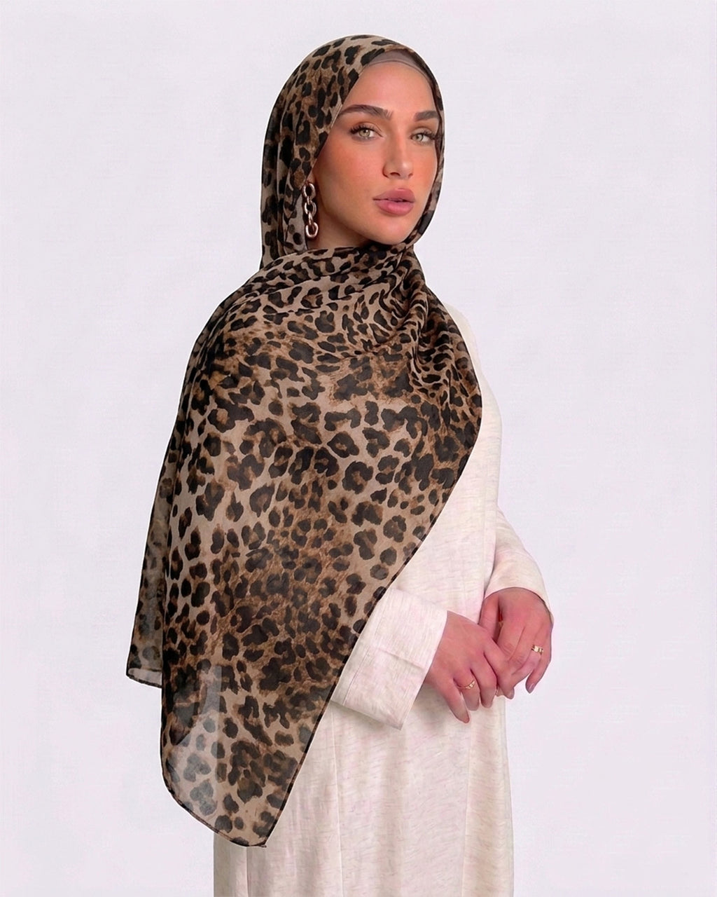 A woman wearing a classic leopard print modal hijab with warm tan, camel, and deep black rosettes, paired with a cream shirt dress.