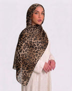 A woman wearing a classic leopard print modal hijab with warm tan, camel, and deep black rosettes, paired with a cream shirt dress.
