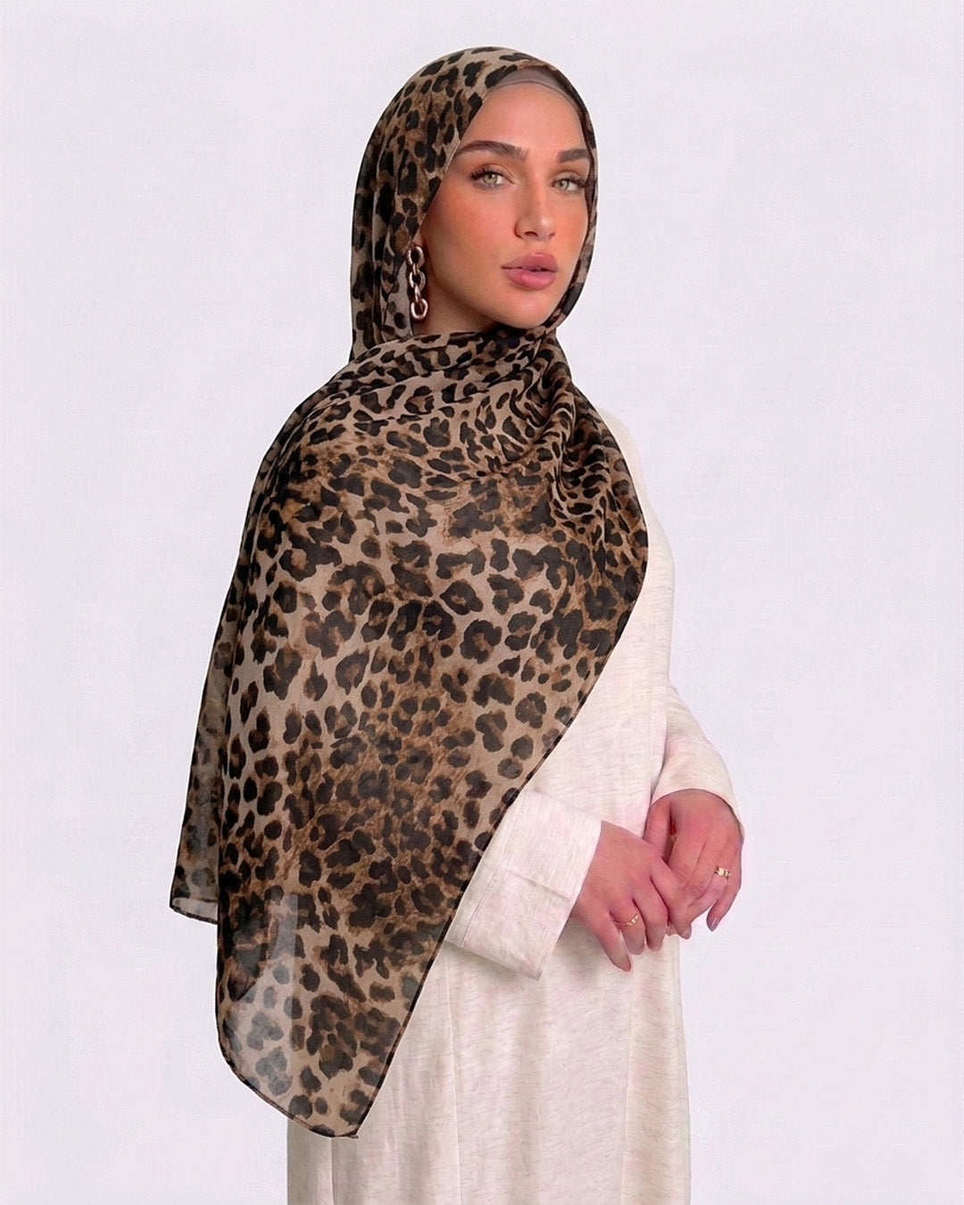 A woman wearing a classic leopard print modal hijab with warm tan, camel, and deep black rosettes, paired with a cream shirt dress.