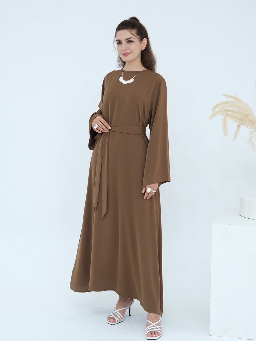 A woman modeling a long-sleeved brown maxi dress with a matching waist tie belt, styled with a white necklace and white strappy heels against a clean white studio background.