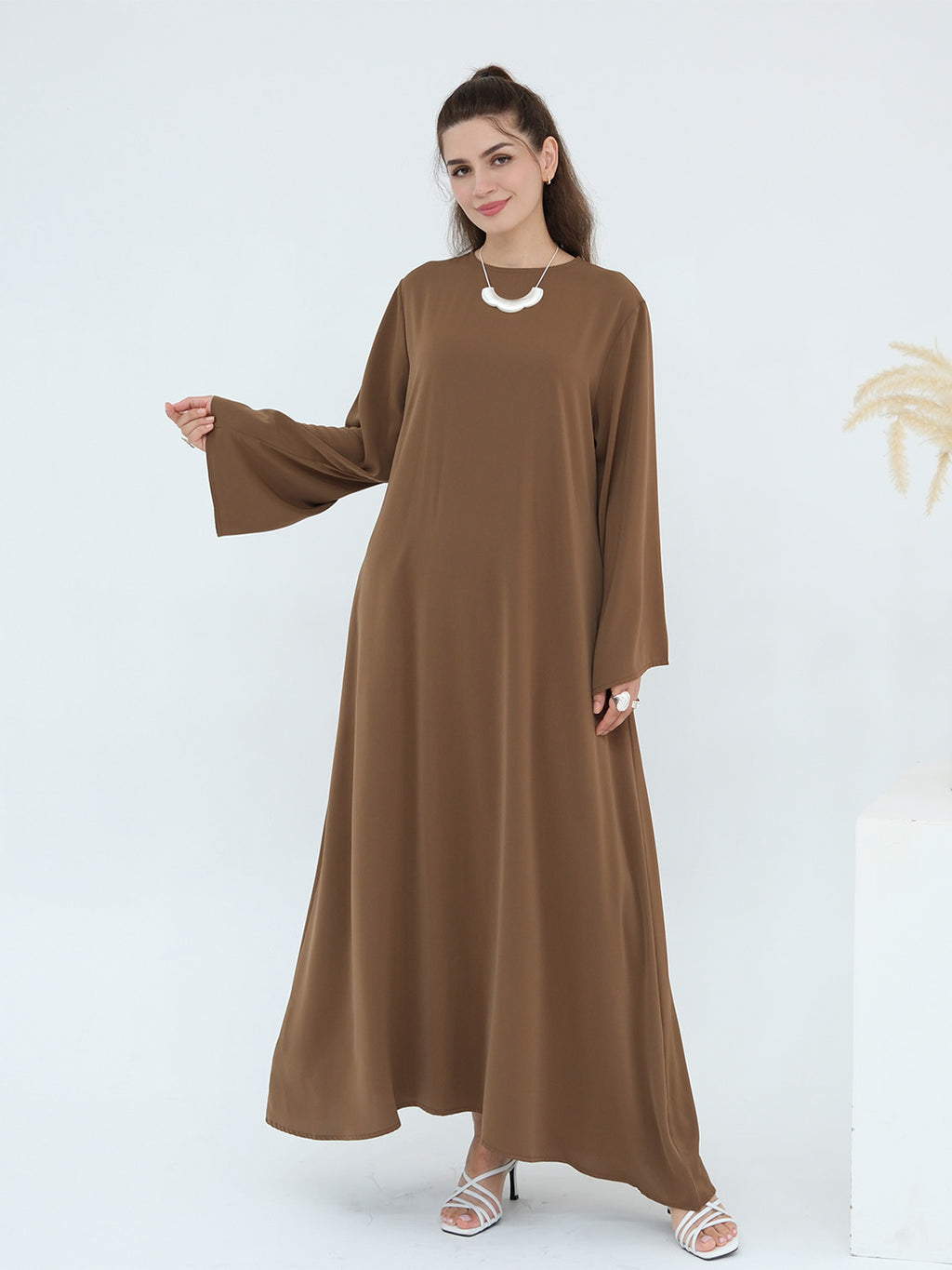 A woman modeling a long-sleeved, floor-length brown abaya dress with a round neckline and a draped silhouette, styled with a white statement necklace and white heeled sandals against a clean white studio background.