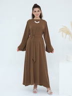 A woman modeling a long-sleeved brown maxi dress with a matching waist tie belt, standing against a clean white studio background.